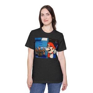 May include: A dark heather grey t-shirt featuring a cartoon image of Mario from the Super Mario Bros. video game series. Mario is looking at three other characters in the background. The text "It's-a me, Mario!" is printed on the shirt.
