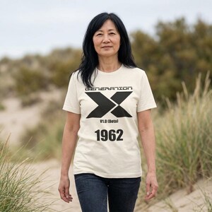 以下が含まれることがあります： クリーム色の半袖Tシャツ。黒いグラフィックデザインが施されており、「GENERATION」の文字の下に大きなスタイライズされた「X」のシンボル、その下に「V1.0 (Beta)」というテキストと「1962」という数字が太い黒のサンセリフ体でプリントされています。