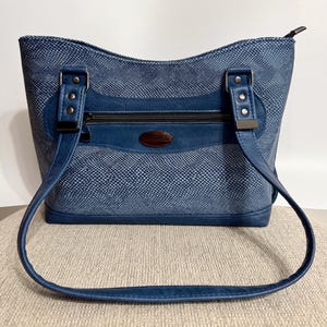 May include: A blue shoulder bag featuring a textured snakeskin-like pattern across the body. The bag includes two blue straps attached with silver-toned rivets, a horizontal zippered front pocket, and a dark oval brand plaque centered on the front.
