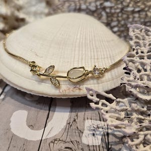 May include: A gold bracelet with a floral design, including a tulip-shaped centrepiece and leaf details. The bracelet is embellished with small crystals. The jewellery is displayed on a seashell.
