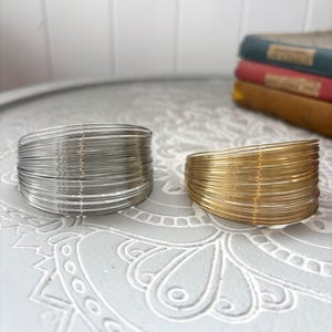 Puede incluir: Dos pulseras de alambre, una plateada y otra dorada, se muestran juntas. Cada pulsera está hecha de numerosos alambres finos y enrollados, creando una apariencia texturizada. Se exhiben sobre una superficie gris con patrones.