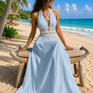 May include: A light blue maxi dress featuring a deep V-neckline, a halter neck strap, and a waistline accented with decorative seashell trim. The dress is crafted from a textured fabric and flows into a long, loose skirt that reaches the ankles.