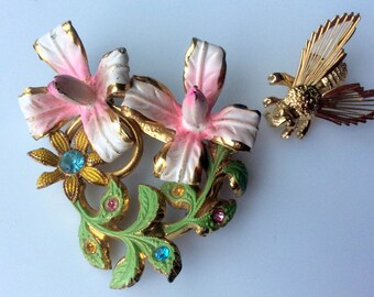 Puede incluir: Un broche de tono dorado con un grupo de flores rosas y blancas con hojas verdes y brillantes piedras de cristal azules y rosas. Un broche de tono dorado separado representa una abeja estilizada con alas intrincadas.