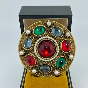 Könnte beinhalten: Goldfarbene Brosche mit kreisförmigem Design, mit einem großen roten Mittelstein, umgeben von kleineren farbigen Steinen und Kunstperlen. Die Brosche wird in einer schwarzen Schatulle mit goldfarbenem Samtinterieur präsentiert, die ihren Vintage-Stil zeigt.