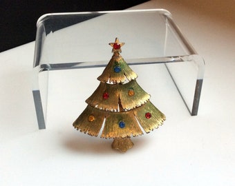 May include: Gold-tone Christmas tree brooch with red, blue, and yellow rhinestone ornaments. The tree has a star on top.