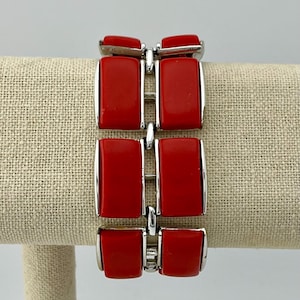 Puede incluir: Una pulsera de eslabones con seis segmentos rectangulares rojos y lisos engastados en metal plateado pulido. El diseño consta de dos columnas paralelas de tres piezas rectangulares conectadas por bisagras de metal plateado, creando una apariencia geométrica estructurada.