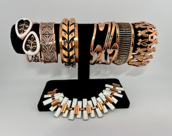 May include: A set of copper and white enamel bracelets and a necklace. The bracelets feature various geometric and floral designs. The necklace is made of copper links and white enamel squares.