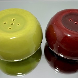 Puede incluir: Un juego de dos saleros y pimenteros de cerámica con forma de manzana. Los saleros son de color verde y rojo con un acabado brillante. Cada salero tiene cinco agujeros en la parte superior para dispensar sal y pimienta.