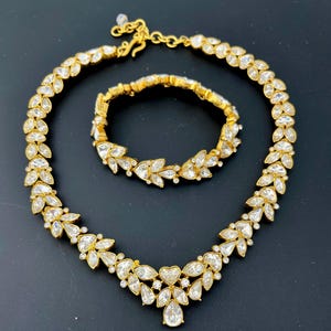 May include: A gold-toned necklace and matching bracelet set featuring a repeating leaf-like pattern composed of clear, marquise-cut crystals. The necklace includes a central heart-shaped crystal and an adjustable chain with a hook clasp closure.