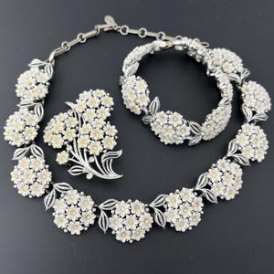 Puede incluir: Un conjunto de joyería en tono plateado que consta de un collar, una pulsera y un broche. Cada pieza presenta racimos de pequeñas flores de color crema con hojas plateadas texturizadas. El collar y la pulsera tienen una estructura de cadena.