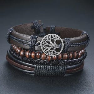 Könnte beinhalten: Mehrschichtiges Armband mit braunem Leder, Holzperlen und geflochtenem schwarzem Kordel. Ein silberfarbener Lebensbaum-Anhänger ist auf dem Armband zentriert. Das Armband ist für eine bequeme Passform konzipiert.