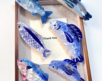May include: A collection of six fish-shaped hair clips in various shades of blue and white. The clips feature translucent, pearlescent, and solid blue acrylic designs with detailed fins, scales, and eyes. Some clips have a spring mechanism.