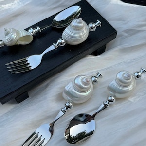 May include: Silverware set featuring forks and spoons with handles adorned with natural seashells. The utensils are displayed on a black surface and a white cloth, showcasing a coastal or nautical theme.