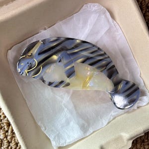 May include: A manatee-shaped hair clip in shades of blue, gray, and yellow. The clip has a glossy finish and is resting on white tissue paper inside a beige container. The manatee design features striped patterns.