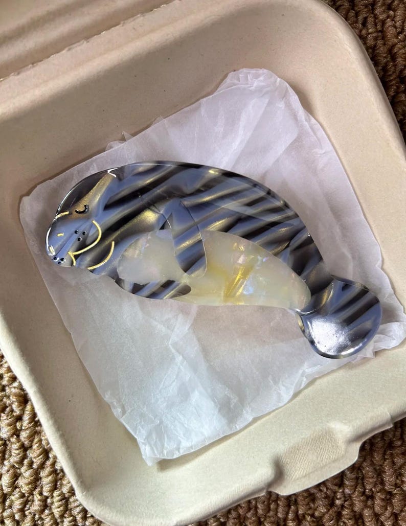 May include: A manatee-shaped hair clip in shades of blue, gray, and yellow. The clip has a glossy finish and is resting on white tissue paper inside a beige container. The manatee design features striped patterns.