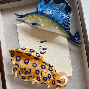 Op de afbeelding: Ingelijst kunstwerk met twee haarclips met marinethema. De ene is een blauw-gele zeilvis, de andere een oranje octopus met blauwe cirkels. Een kleine kaart met de tekst "Have a nice day" is ook inbegrepen.