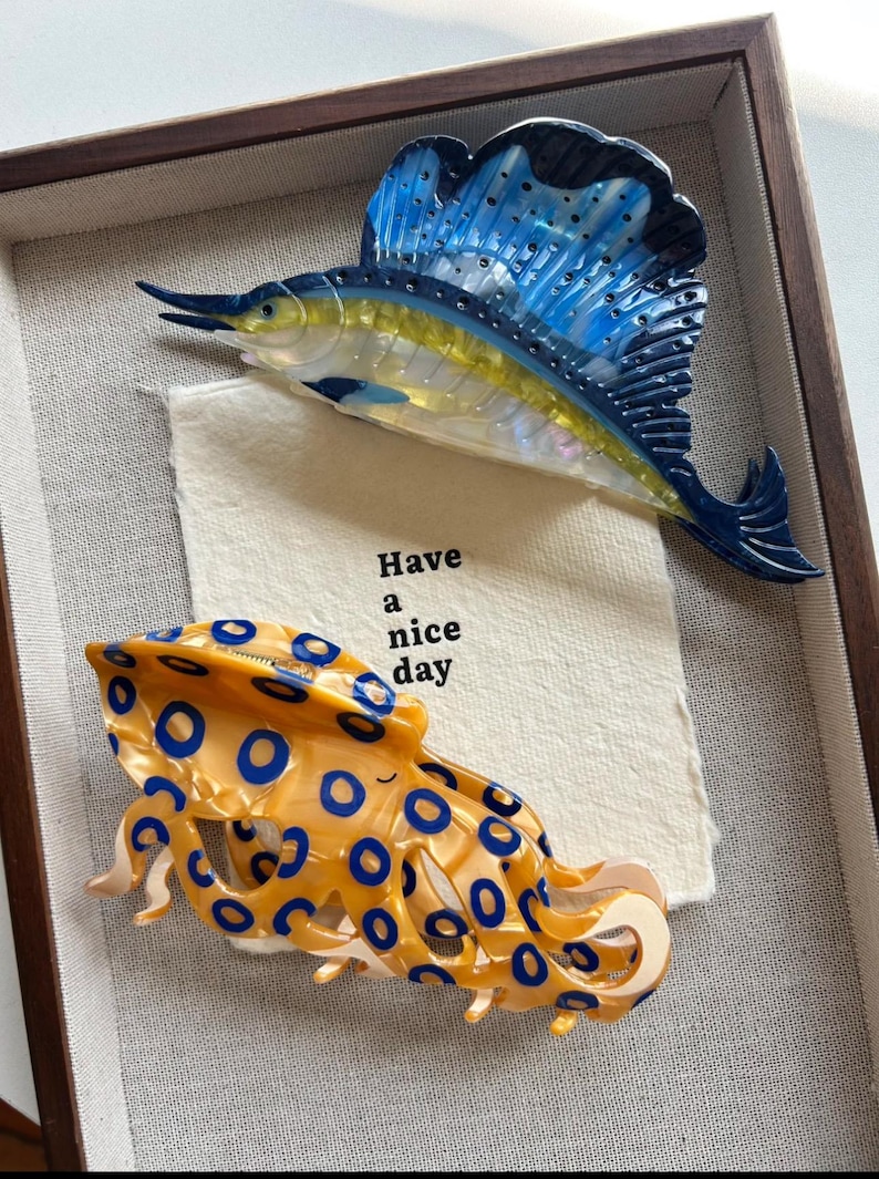 May include: Framed artwork featuring two marine-themed hair clips. One clip is a blue and yellow sailfish, the other is an orange octopus with blue circles. A small card with the text "Have a nice day" is also included.