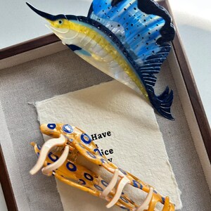 May include: A decorative fish-shaped hair clip and a fish-shaped brooch. The brooch is a sailfish with blue, yellow, and black accents. The hair clip is yellow with blue circles and cream-colored accents. The text "Have a nice" is visible.