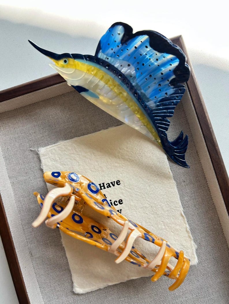 May include: A decorative fish-shaped hair clip and a fish-shaped brooch. The brooch is a sailfish with blue, yellow, and black accents. The hair clip is yellow with blue circles and cream-colored accents. The text "Have a nice" is visible.