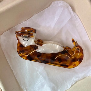 May include: A brown and tan otter-shaped hair clip. The clip is made of a tortoiseshell-patterned material with a white belly and a smiling face. The otter is lying on its back. The hair accessory is on white tissue paper.