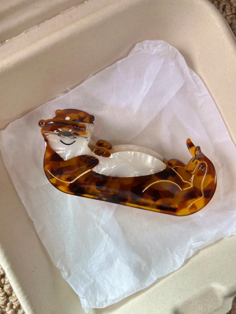 May include: A brown and tan otter-shaped hair clip. The clip is made of a tortoiseshell-patterned material with a white belly and a smiling face. The otter is lying on its back. The hair accessory is on white tissue paper.