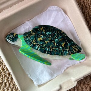 May include: A decorative sea turtle figurine with a green and gold shell, and green flippers. The figurine is resting on white tissue paper inside a cardboard container. The turtle's shell has a marbled appearance.