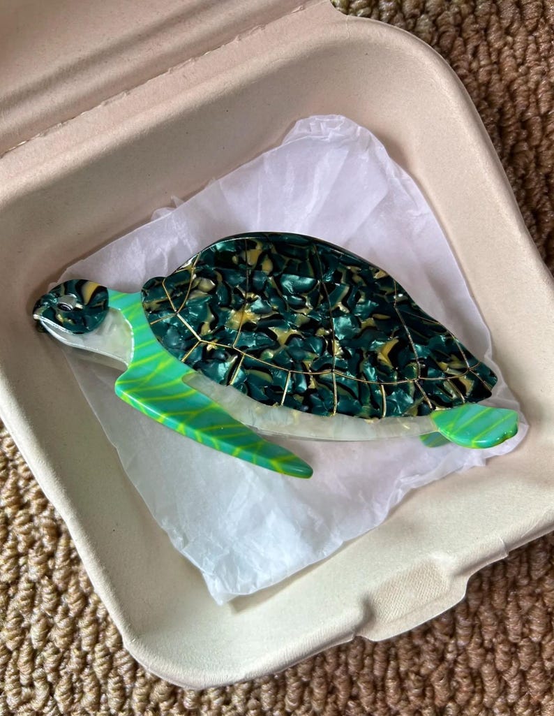 May include: A decorative sea turtle figurine with a green and gold shell, and green flippers. The figurine is resting on white tissue paper inside a cardboard container. The turtle's shell has a marbled appearance.