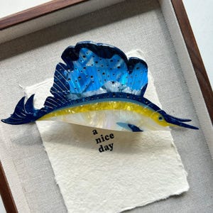 May include: A framed artwork featuring a stylized sailfish. The fish has a blue and white sail, a yellow body, and a long, pointed snout. The artwork includes the text "a nice day" on a textured paper background, set within a wooden frame.