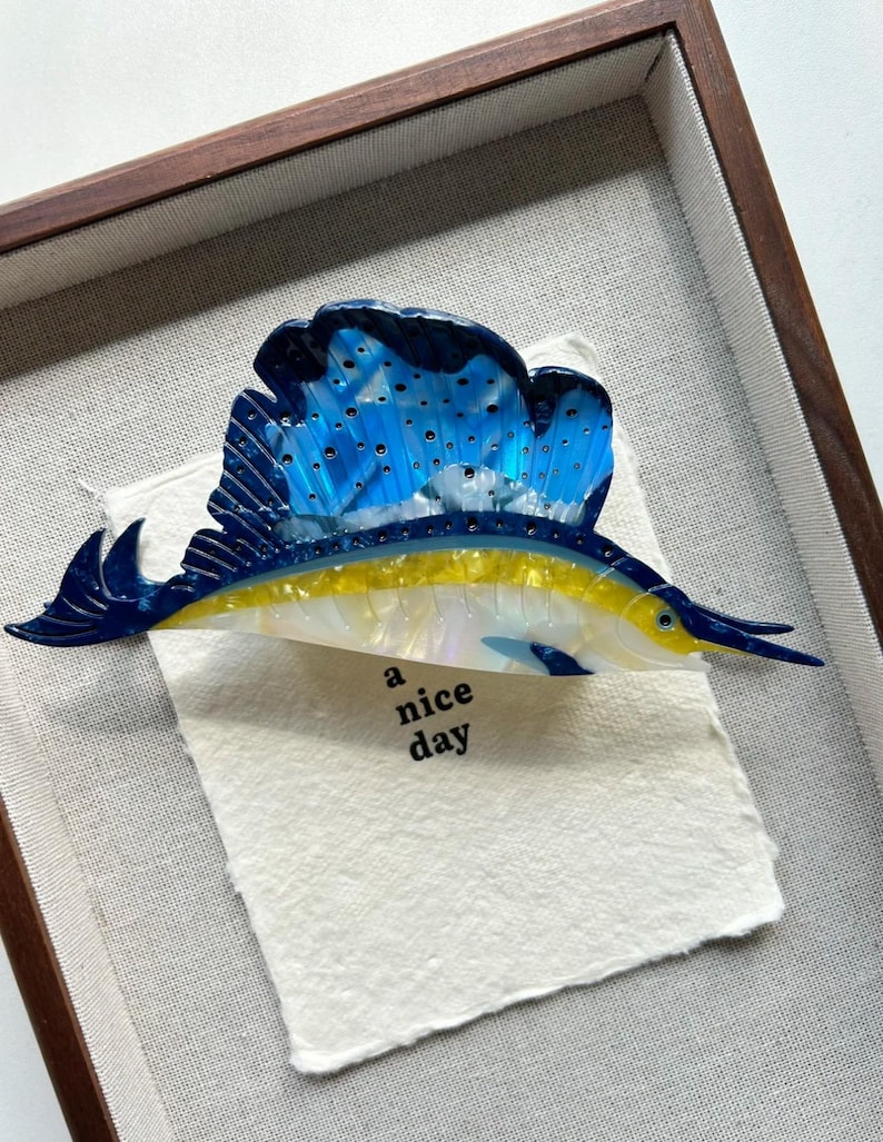 May include: A framed artwork featuring a stylized sailfish. The fish has a blue and white sail, a yellow body, and a long, pointed snout. The artwork includes the text "a nice day" on a textured paper background, set within a wooden frame.