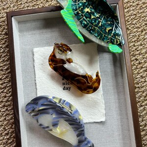May include: Three decorative hair clips in a shadow box. The clips are shaped like a sea turtle, an otter, and a manatee. The sea turtle clip is green and black. The otter clip is brown and white. The manatee clip is blue and white.