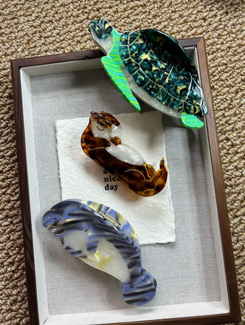 May include: Three decorative hair clips in a shadow box. The clips are shaped like a sea turtle, an otter, and a manatee. The sea turtle clip is green and black. The otter clip is brown and white. The manatee clip is blue and white.