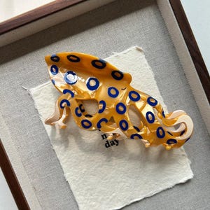 May include: A decorative hair clip in the shape of an octopus. The clip is a vibrant yellow with blue circular accents. It rests on a white fabric inside a wooden frame. The word "day" is visible on the fabric.