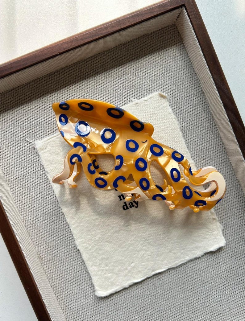 May include: A decorative hair clip in the shape of an octopus. The clip is a vibrant yellow with blue circular accents. It rests on a white fabric inside a wooden frame. The word "day" is visible on the fabric.