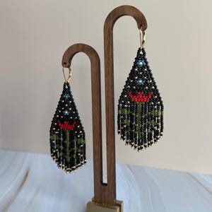 May include: Beaded dangle earrings with a black background, featuring a red flower and green stems. The earrings have gold-colored accents and are displayed on a wooden stand. The earrings are approximately 3 inches long.