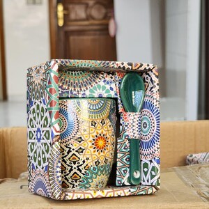May include: A decorative ceramic mug and spoon set. The mug and spoon are green and feature a colorful geometric pattern. The mug is packaged in a box with a matching design, making it a great gift.