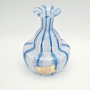 May include: A clear glass vase with vertical blue stripes and an internal white latticino mesh pattern. The vase has a bulbous body and a flared rim. A rectangular paper label is attached to the front with the text INDUSTRIE VENEZIANE DI B. BATTISTON S. MARCO, 1320 VENEZIA MURANO GLASS MADE IN ITALY.
