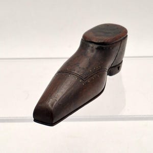 May include: A small, dark brown wooden object shaped like a shoe with a square toe and a low heel. The surface features decorative rows of small, evenly spaced metal studs along the vamp and collar, creating a detailed, structured appearance.