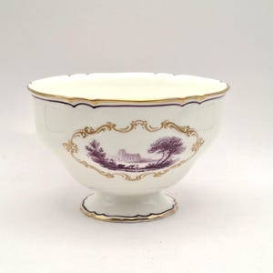 May include: A white porcelain bowl with a scalloped rim and a pedestal base. The exterior features a central purple landscape illustration of a building and trees, framed by an ornate gold border. The rim and base are accented with gold trim.