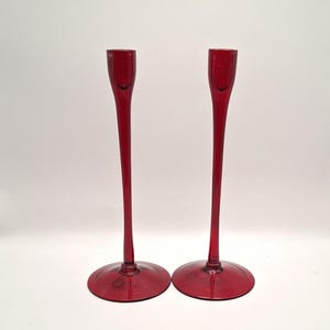 May include: A pair of elegant red glass candle holders. They feature a circular base, a long, thin stem, and a small cup to hold a candle. The candle holders are a deep red hue and are set against a plain white backdrop.