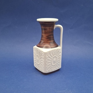 May include: A decorative ceramic vase with a white handle and a square base. The vase features a brown, wood-grain patterned neck and a white, embossed floral design on the body. The vase is approximately 18 cm tall.
