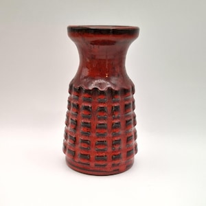 May include: A ceramic vase featuring a vibrant red and dark brown glaze. The body of the vase has a textured, grid-like pattern of raised squares. It has a wide, flared rim and a cylindrical neck that tapers down into a wider, structured base.