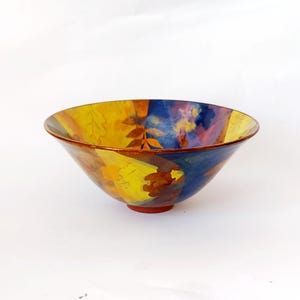 May include: A decorative ceramic bowl with a colourful leaf design. The bowl features a vibrant mix of yellow, blue, and orange hues, with a reddish-brown rim and base. The bowl's interior is adorned with leaf patterns.