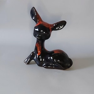 May include: A ceramic deer ornament in a resting pose. The deer has a glossy finish with a colour scheme of black and reddish-brown. The ornament's ears are upright, and its body is in a relaxed, seated position. This decorative item is suitable for home decor.