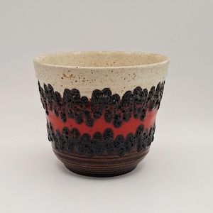 May include: A ceramic cup featuring a speckled cream-coloured rim, a central band of vibrant red glaze, and a textured, porous black lava-like finish. The lower portion of the cup displays a dark brown, horizontally ribbed surface with a rounded base.