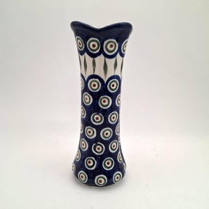 May include: A ceramic vase with a flared, wavy rim and a tapered body. The surface features a repeating pattern of dark blue, light green, and brown concentric circles on a white background, with vertical green leaf-like accents near the top.