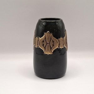 May include: A black ceramic vase with a gold-coloured decorative band. The vase has a cylindrical shape with a slightly flared top. The gold band features a repeating floral design. The vase is likely used for decorative purposes.