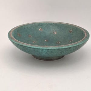 May include: A shallow, round teal bowl with a mottled surface texture. The interior features small, light-coloured floral patterns scattered across the base, while the rim is decorated with a scalloped border in a muted, metallic-toned finish.