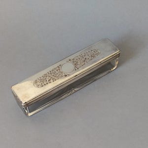 May include: A rectangular, silver-coloured box with a clear glass base. The lid features ornate, symmetrical floral and scrollwork designs. The box is likely a vintage or antique item, possibly for storing small items or jewellery.