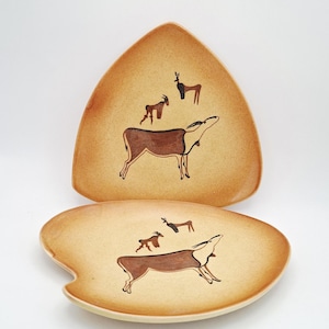 May include: Two ceramic plates with a speckled tan finish, featuring hand-painted brown and black illustrations of antelopes. One plate is triangular in shape, while the other is an irregular oval with a curved indentation on the left side.