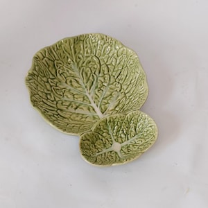 May include: Two ceramic serving dishes shaped like cabbage leaves. The larger dish is round, and the smaller is oval. Both are light green with detailed veining.